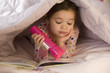 Mixed race girl reading book in bed with flashlight