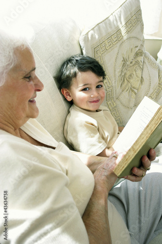 Photo: An old woman sitting on the couch reading book while her grandson is sitting beside her An old woman sitting on the couch reading book while her grandson is sitting beside her