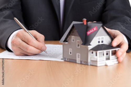 Photo: Businessman signs contract behind house architectural model Businessman signs contract behind house architectural model