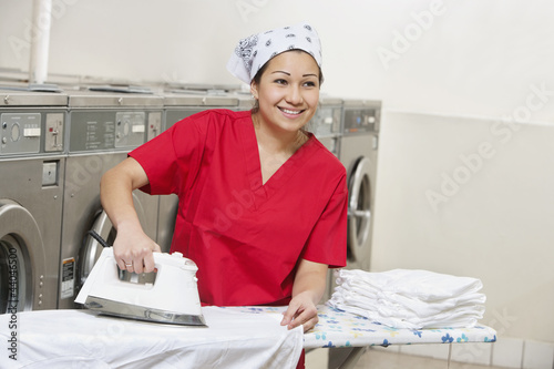Photo: Happy young employee wearing bandana while ironing in Laundromat Happy young employee wearing bandana while ironing in Laundromat