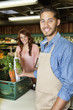 Portrait of a happy young sales clerk holding vegetables with woman in background