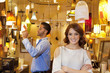 Portrait of young woman with arms crossed while man looking at price tag in background in lights store
