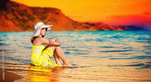 Photo: beautiful girl with hat sitting on the beach in a yellow dress beautiful girl with hat sitting on the beach in a yellow dress