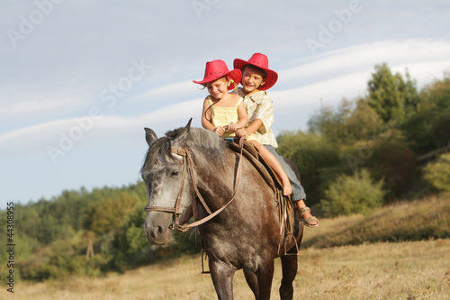 Photo: two happy children in cowboy hats riding horse on natural backgr two happy children in cowboy hats riding horse on natural backgr