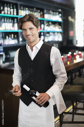 Photo: Portrait of smiling sommelier holding bottle of wine in bar Portrait of smiling sommelier holding bottle of wine in bar