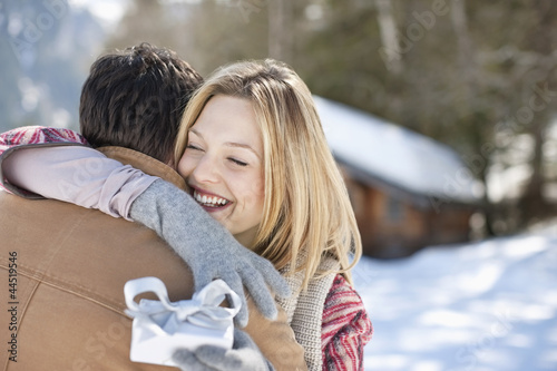 Photo: Smiling woman holding Christmas gift and hugging man in snowy field Smiling woman holding Christmas gift and hugging man in snowy field