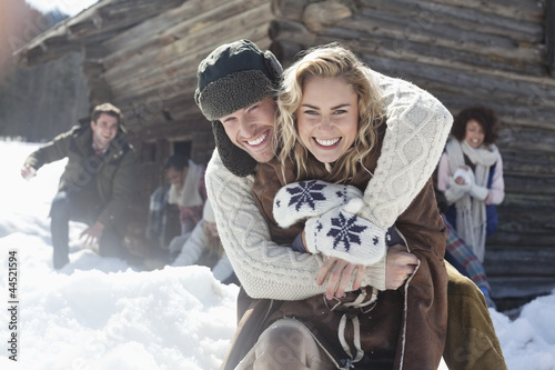 Photo: Portrait of smiling couple hugging with friends in background Portrait of smiling couple hugging with friends in background