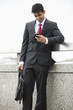 Photo: Young Indian businessman texting through cell phone while holding laptop bag at parapet Young Indian businessman texting through cell phone while holding laptop bag at parapet