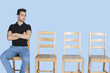 Photo: Portrait of a young man sitting beside empty wooden chairs over blue background Portrait of a young man sitting beside empty wooden chairs over blue background