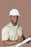 Photo: Portrait of happy young African male construction worker holding clipboard over brown background Portrait of happy young African male construction worker holding clipboard over brown background