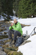 Smiling man with backpack and ski poles sitting at edge of stream in snowy woods