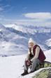 Portrait of smiling senior woman sitting on rock on snowy mountain