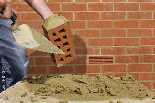 Photo: Close up of bricklayer applying mortar to brick with trowel in front of wall Close up of bricklayer applying mortar to brick with trowel in front of wall