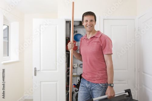 Photo: Portrait of smiling handyman holding toolbox and copper pipes in front of boiler in closet Portrait of smiling handyman holding toolbox and copper pipes in front of boiler in closet