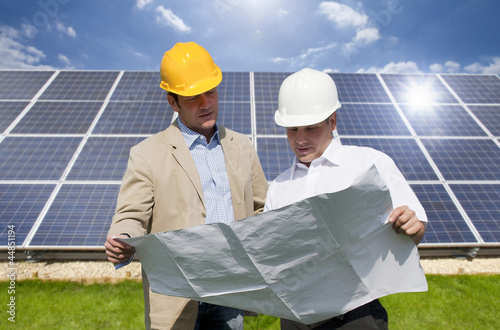 Photo: Technicians holding blueprints talking near large solar panels Technicians holding blueprints talking near large solar panels