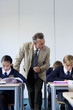Photo: Male teacher looking over students taking exam at desk in classroom Male teacher looking over students taking exam at desk in classroom