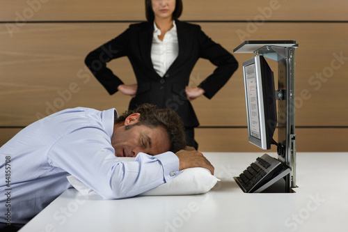 Photo: Angry businesswoman looking down at businessman sleeping on pillow at desk Angry businesswoman looking down at businessman sleeping on pillow at desk