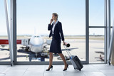 Photo : Businesswoman in suit pulling suitcase and talking on cell phone at airport window Businesswoman in suit pulling suitcase and talking on cell phone at airport window