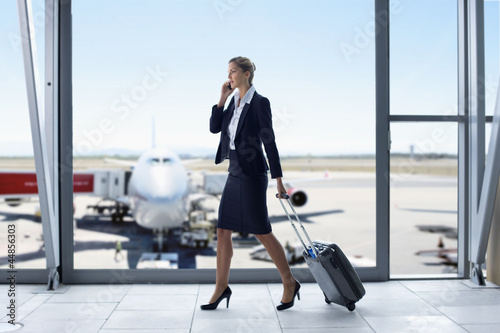 Photo: Businesswoman in suit pulling suitcase and talking on cell phone at airport window Businesswoman in suit pulling suitcase and talking on cell phone at airport window