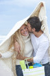 Portrait of smiling young couple with blanket and basket kissing under blanket on beach