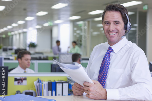 Photo: Portrait of smiling businessman wearing headset and reviewing report in office Portrait of smiling businessman wearing headset and reviewing report in office