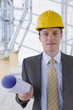 Photo: Portrait of smiling architect wearing suit and hard-hat and holding blueprints in modern lobby Portrait of smiling architect wearing suit and hard-hat and holding blueprints in modern lobby