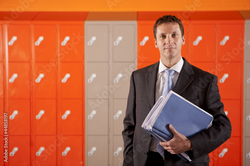 Photo: Serious teacher holding paperwork near school lockers Serious teacher holding paperwork near school lockers