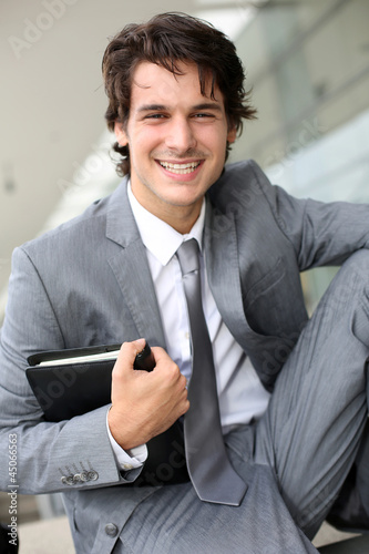 Photo: Smiling young businessman sitting outside building Smiling young businessman sitting outside building