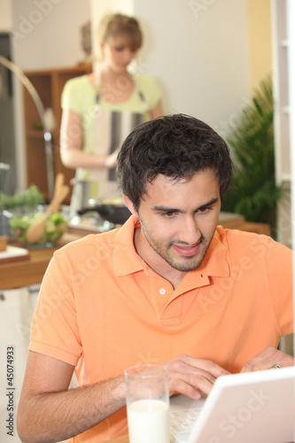 Photo: Man looking at his laptop while his wife prepares dinner Man looking at his laptop while his wife prepares dinner