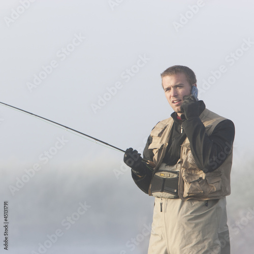 Photo: man fishing in a river on his cell phone man fishing in a river on his cell phone