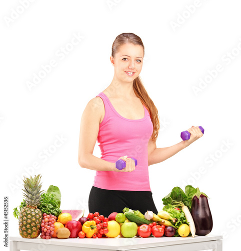 Photo: Smiling woman holding weights and table full of fruits and veget Smiling woman holding weights and table full of fruits and veget
