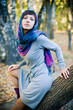 The girl in a scarf and in a knitted dress sits on a tree.
