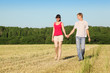 Husband, wife holding hands walk in field near wood