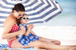 Mother And Daughter Under Beach Umbrella Putting On Sun Cream