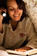 Smiling Woman Reading a Book with Flashlight on Bed