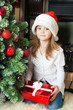 Little girl in Santa hat with gift near christmas tree