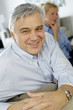 Smiling senior man sitting in couch, wife in background