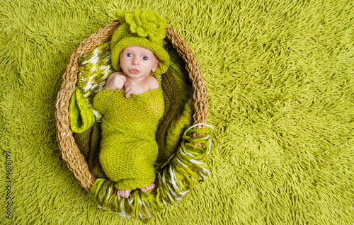 Photo: Newborn baby in woolen hat inside basket over green background Newborn baby in woolen hat inside basket over green background