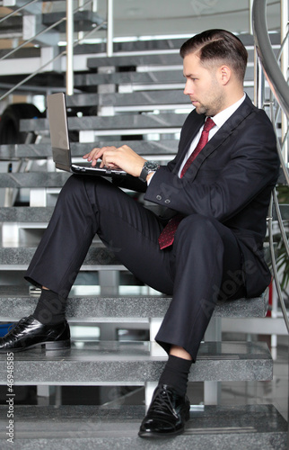 Photo: Handsome businessman sitting on the stairs and using his laptop Handsome businessman sitting on the stairs and using his laptop