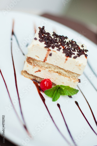Photo: Tiramisu cake with a cherry and mint on white plate on table in Tiramisu cake with a cherry and mint on white plate on table in