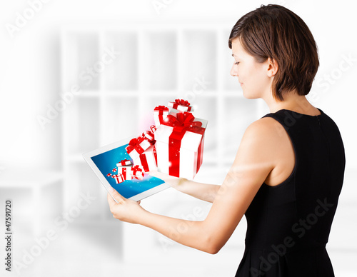 Photo: Young woman looking at modern tablet with present boxes Young woman looking at modern tablet with present boxes