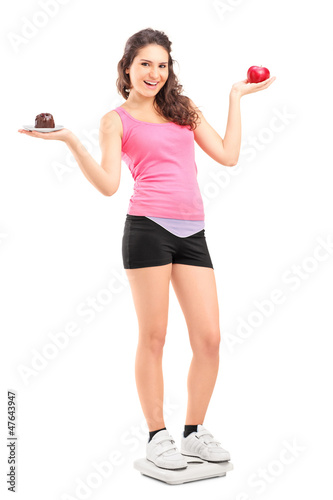 Photo: Smiling female on a weight scale holding a red apple and cake Smiling female on a weight scale holding a red apple and cake