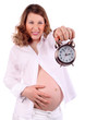 Dissatisfied pregnant woman in white holds at arm alarm clock