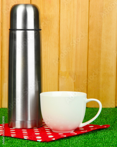 Photo: metal thermos with cup on grass on wooden background metal thermos with cup on grass on wooden background