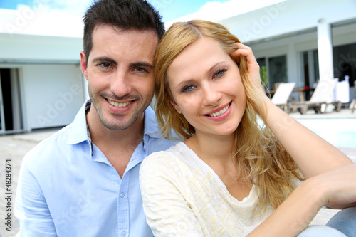 Photo: Cheerful young couple sitting in front of modern house Cheerful young couple sitting in front of modern house