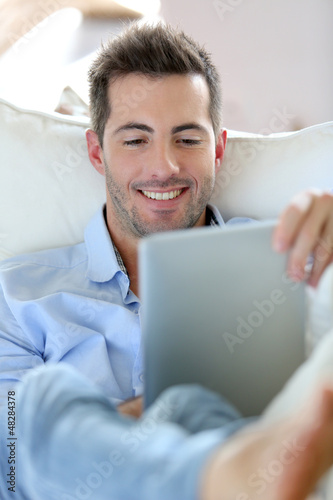 Photo: Cheerful young man laying in sofa with digital tablet Cheerful young man laying in sofa with digital tablet