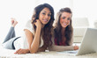 Two smiling women typing on a computer while lying on the floor