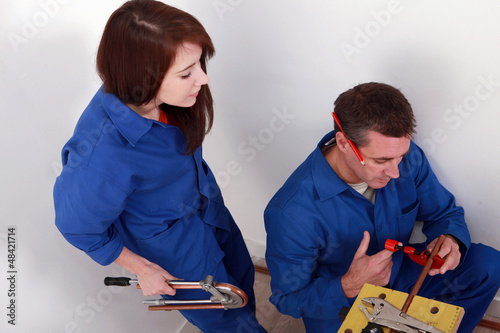 Photo: Plumber cutting copper pipe while his female apprentice watches Plumber cutting copper pipe while his female apprentice watches