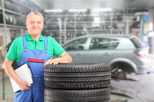 Photo: Mature mechanic posing on a tires in front of car at repair shop Mature mechanic posing on a tires in front of car at repair shop