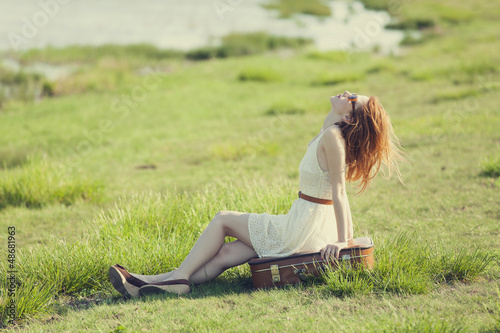 Photo: Young fashion girl with suitcase sitting at spring grass near la Young fashion girl with suitcase sitting at spring grass near la